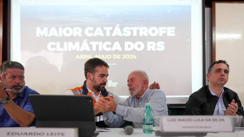 Foto da entrevista coletiva, na qual Leite e Lula aparecem sentados junto a uma mesa. Eles olham um para o outro e se dão as mãos. Lira, à esquerda, e Pacheco, à direita, aplaudem. Ao fundo, há uma apresentação de slide na qual está escrita "Maior catástrofe climática do RS".
