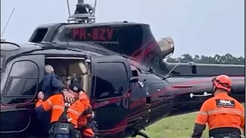Foto de uma pessoa resgatada entrando num helicóptero, amparada por dois profissionais.