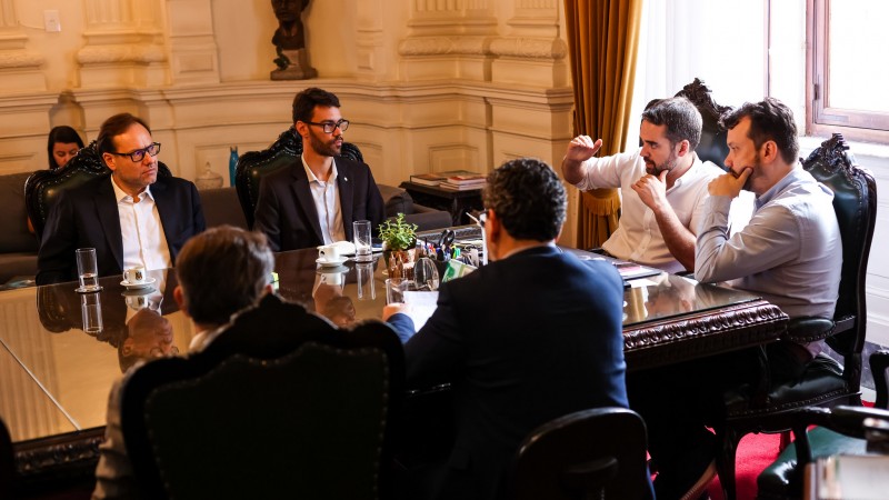 Em torno de uma mesa de reuniões, onde estão quatro homens de camisa e paletó, o governador Eduardo Leite, de camisa branca, e o chefe da Casa Civil, Artur Lemos, de camisa azul, estão sentados na cabeceira, à direita da imagem.