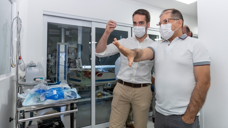 Eduardo Leite, de camisa branca e cal&ccedil;a bege, em p&eacute;, gesticula com a m&atilde;o direita ao lado de um homem de camisa polo branca e cal&ccedil;a jeans, que sinaliza para &aacute;rea diante de si, em uma sala com equipamentos hospitalares, durante visita ao Hospital de Caridade de Carazinho. Ambos usam m&aacute;scara.