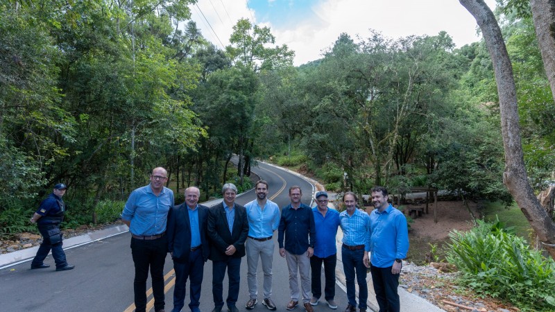 O governador Eduardo Leite acompanhado de comitiva posa para foto na via, agora pavimentada, que dá acesso ao Parque Olivas de Gramado, na Serra. Ao fundo, arvores que margeiam o traçado da estrada.