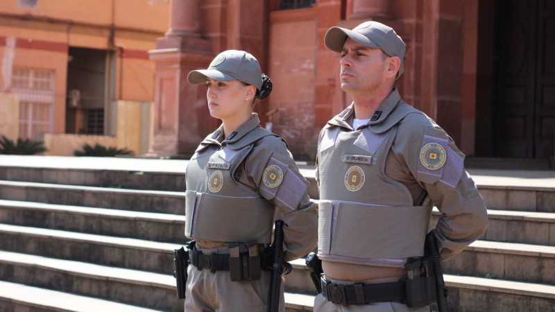 Dois policiais militares, sendo uma mulher e um homem, aparecem em pé, lado a lado, fardados, com os braços em posição descansar junto às costas, diante da escadaria de um prédio.