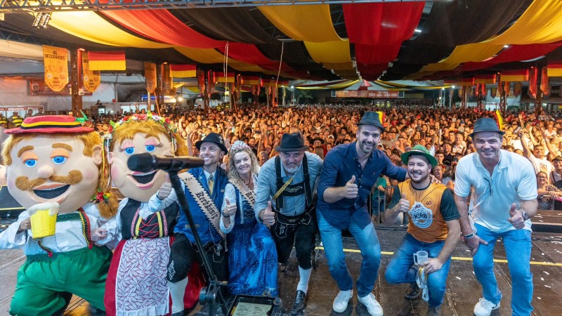 O governador Eduardo Leite posa para foto ao lado da corte do 40º Festival do Kerb em Estância Velha. Ele está de calça jeans, camisa azul escura e com um chapé preto. Há pessoas com trajes típicos alemães e dois bonecos, representando um casal típico de imigrantes. Ao fundo, um espaço lotado pelo público do evento, sob um cobertura de tecidos nas cores preto, vermelho e amarelo, da bandeira alemã.