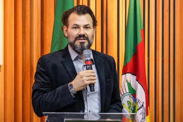 Diretor-executivo do RS, Antônio Padilha, fala segurando um microfone. Ele está de casaco preto e camisa lilás claro. Ao fundo, parede com ripas de madeira e bandeiras do RS e do Brasil.
