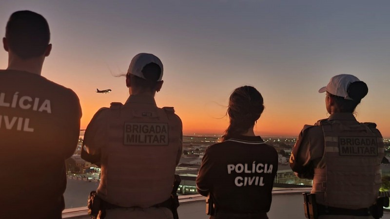 A foto mostra quatro policiais de costas em uma saca observando o horizonte da cidade no crepúsculo, com uma avião voando ao fundo. Da esquerda pra direita, um homem com camiseta da Polícia Civil, uma mulher com colete da BM, uma mulher com camiseta da PC, e uma outra mulher com colete da BM.