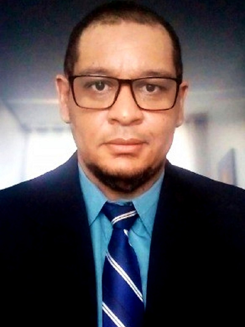 Denilson está de terno preto, camisa azul e gravata azul, e usa óculos