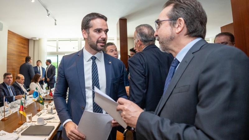 Leite, ao centro, conversa com outra pessoa numa sala em que se vê uma mesa ao fundo, na qual políticos, secretários e diretores estão sentados