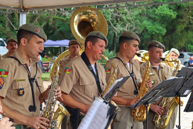 Quatros policiais militares da banda da Brigada Militar, todos fardados, tocam saxofones diante de pranchetas com partituras. Ao fundo, outros policiais músicos tocam outros instrumentos de sopro.
