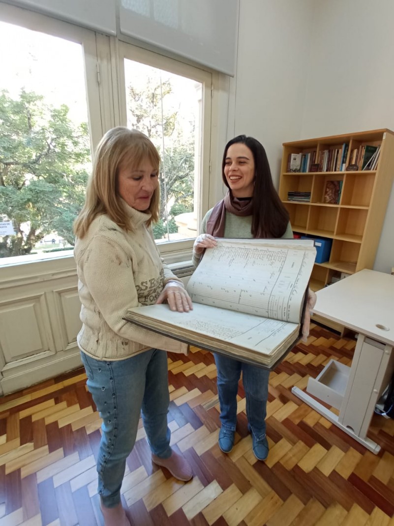 Numa sala fechada, as duas mulheres seguram o documento, que se assemelha a um grande livro. A da esquerda possui cabelos longos e loiros e usa su&eacute;ter branco; a da direita tem cabelos longos e escuros, e sorri