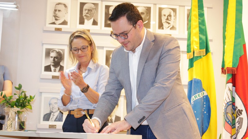 Wesp assina um documento. Ele tem cabelo escuro e curto, usa óculos e blazer cinza. Ao fundo, está uma mulher loira aplaudindo o ato, que acontece numa sala fechada em que estão também as bandeiras do Brasil e do RS