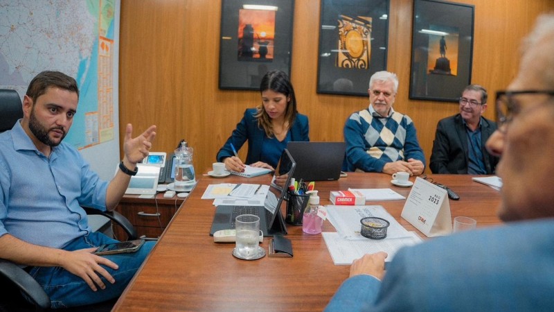 Gabriel Souza aparece no canto esquerdo da foto, sentado atrás de uma mesa. Ele conversa com alguém cujo perfil aparece do lado direito da foto. De frente, na imagem, estão três pessoas que também participam da reunião.