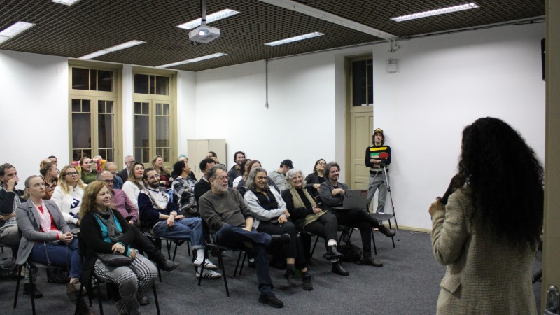 Numa sala fechada, um grupo de pessoas sentadas em classes assiste a uma mulher que fala ao microfone. O grupo é composto por em torno de 20 pessoas, vestidas com roupas de frio.