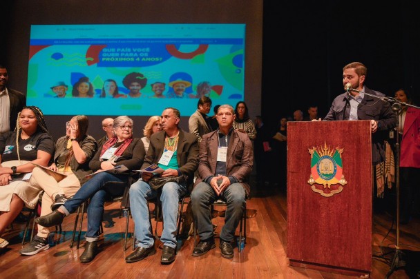 À direita da imagem, no palco no Teatro Barone da ALRS, Gabriel Souza fala ao microfone diante de um púlpito de madeira com o Brasão de Armas do RS. Do centro para esquerda, pessoas participantes do evento sentadas lado a lado em cadeiras. Ao fundo, uma apresentação em um telão.