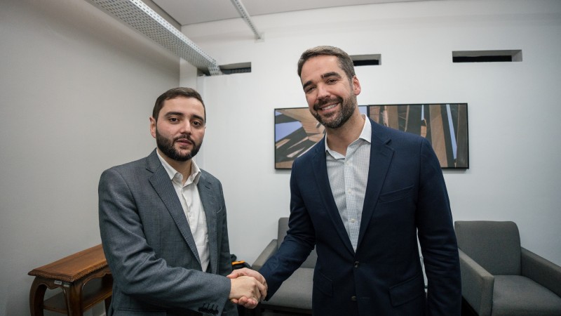 O governador Eduardo Leite, de casa azul marinho e camisa branca, e o vice-governador Gabriel Souza, de casaco cinza e camisa branca, apertam as m&atilde;o. Ao fundo, poltronas cinzas e um quadro com uma fotografia na parede. 