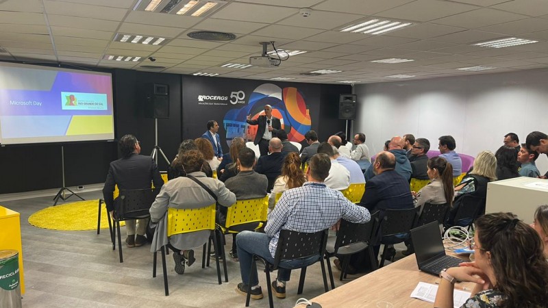 Numa sala fechada, várias pessoas estão sentadas em cadeiras assistindo a um homem que, de pé e ao microfone, realiza a conferência. Ao fundo, está um painel preto onde está escrito "Procergs 50 anos". No slide, a logo do governo e o nome do evento "Microsoft day".