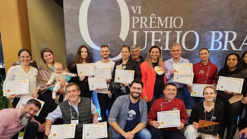 Várias pessoas exibindo os certificados dos queijos premiados no concurso. Formam duas fileiras (um grupo está em pé e outro agachado) à frente de um painel com o nome do concurso Prêmio Queijo Brasil 2023.