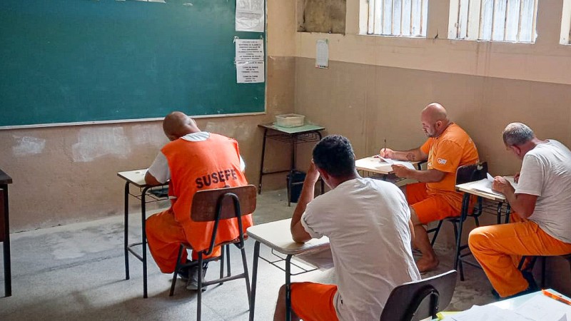 Quatro homens privados de liberdade est&atilde;o sentados em classes no canto direito frontal de uma sala de aula. Eles usam uniformes laranja e branco. &Agrave; frente deles, est&aacute; posicionado um quadro negro. 