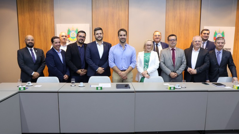 Numa sala de reuni&otilde;es, Eduardo Leite est&aacute; de p&eacute;, ao centro, tendo a seu lado a secret&aacute;ria da sa&uacute;de, Arita, e do turismo, Vilson Covatti. Ao lado deles, h&aacute; v&aacute;rios outros homens tamb&eacute;m de p&eacute;, trajando terno. Todos posam para a foto.