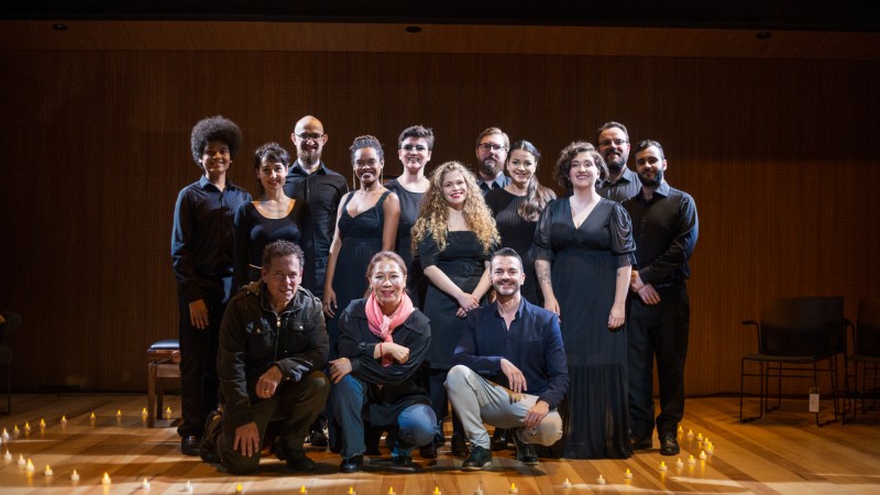 Foto frontal de um grupo de pessoas trajando roupas escuras e que está, em sua maioria, de pé (há três agachados, à frente) no palco do teatro. Eles sorriem para a foto, e a iluminação é levemente amarelada.