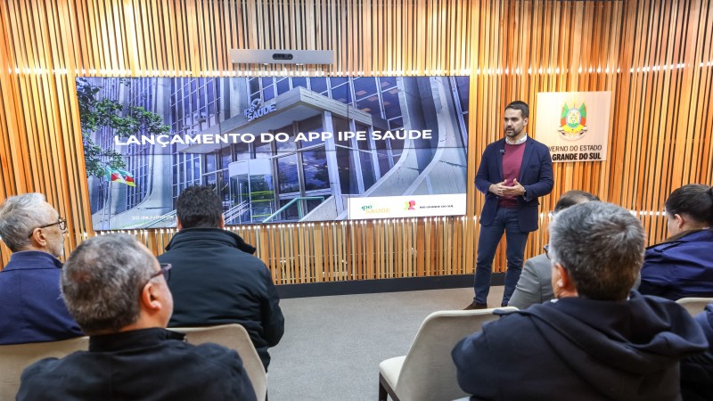 O governador Eduardo leite está em pé diante de uma plateia apresentando o Aplicativo do IPE Sáude. Atrás dele à esquerda há uma tela com o texto: Lançamento do app do IPE Saúde; além de uma placa com o brasão do Rio Grande do Sul mais à direita.