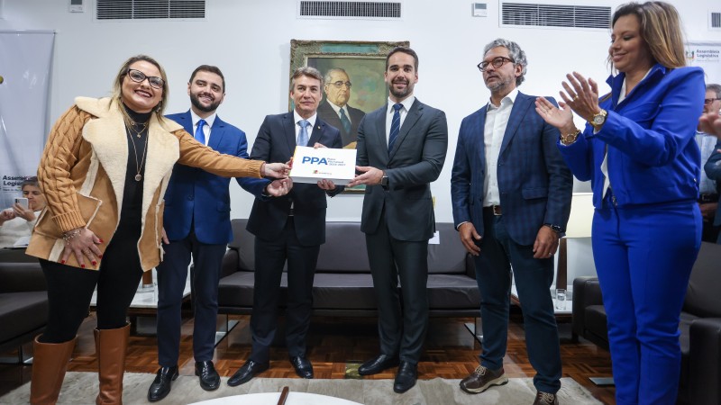 Em uma sala fechada, governador e comitiva posam para a foto segurando o PPA 2024-2027. No centro do grupo, estão o governador e o presidente da Assembleia Legislativa com o documento. 