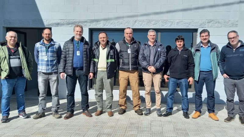 Participantes do encontro posam, de pé, para a foto. São nove homens vestindo roupas de frio, enfileirados de lado. Eles estão ao ar livre, em frente à entrada de um prédio. Estão no sol. 