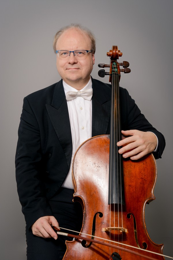 Bernhard Lörcher está de fraque, sentado, segurando seu violoncelo. Ele sorri para a foto. O fundo é cinza. 