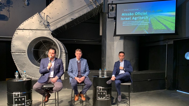 Sentados em bancos altos, três homens estão apresentando um slide no qual se lê Missão Oficial Israel Agritech. Os três trajam ternos, e um deles está com o microfone na mão. Há latões de cor preta que servem como apoio para os copos e garrafas de água.