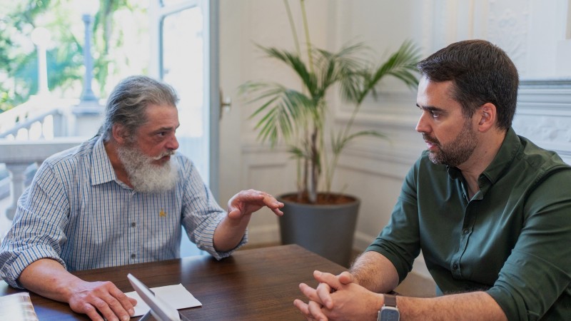 Sentados à mesa, Luiz Marenco à esquerda e Eduardo Leite mais à direita da imagem. Luiz fala e gesticula, Leite escuta atento com os dedos cruzados e apoiados sobre a mesa.