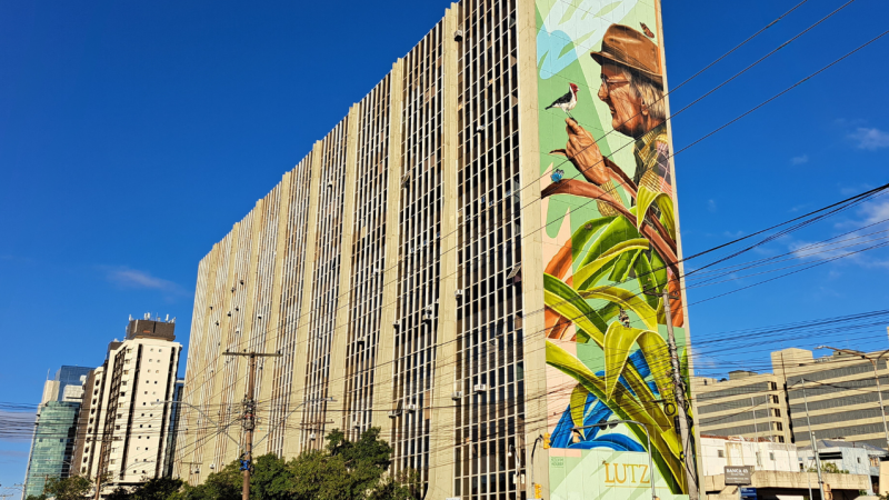 Imagem da fachada do IPE Saúde, mostrando um enorme painel em uma de suas laterais. O painel é uma criação em grafite do artista Kelvin Koubik, muralista natural de Porto Alegre e mostra a imagem de perfil do ambientalista José Lutzenberger de perfil, atrás de umas folhagens, com um passarinho posado em sua mão.