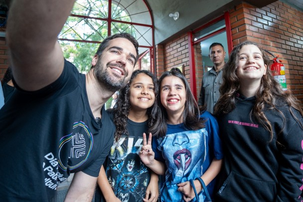 O governador Eduardo Leite, vestindo uma camiseta preta com a logo do Dia do Patrimônio, tira uma selfie com três crianças de cabelo comprido durante a visita à Biblioteca Pública Romano Rief, no bairro IAPI.