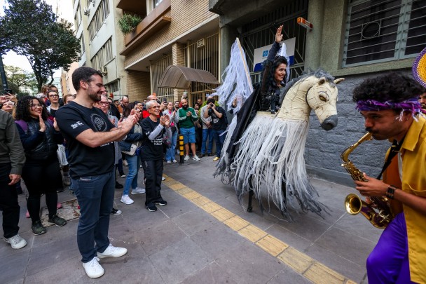 O governador Eduardo Leite e a secretária da Cultura, Beatriz Araújo, ambos vestindo camisetas pretas com a logo do Dia do Patrimônio, aplaudem artista que realiza uma performance em rua do centro de Porto Alegre, montando um cavalo de papelão À direita da imagem, um homem de camisa amarela toca saxofone.