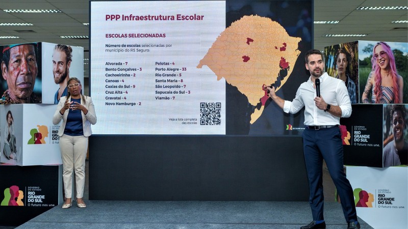 A imagem mostra o governador em pé em um pequeno tablado com microfone na mão apresentando a PPP Infraestrutura Escolar ele aponta coma outra mão para uma tela atrás de si. Ele está do lado direito da imagem. Do lado esquerdo há uma intérprete de libras. Nos extremos da foto, cubos com imagens de pessoas e a logomarca do governo do Estado.