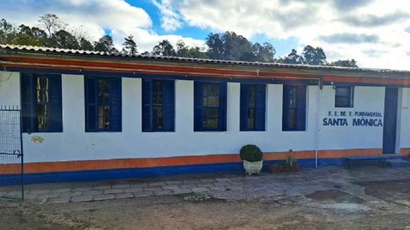 Imagem da fachada da Escola Santa Mônica em Garibaldi. A foto mostra o único piso da escola e d esquerda para direita são vistas seis janelas azuis, um vitrô azul e uma porta azul.