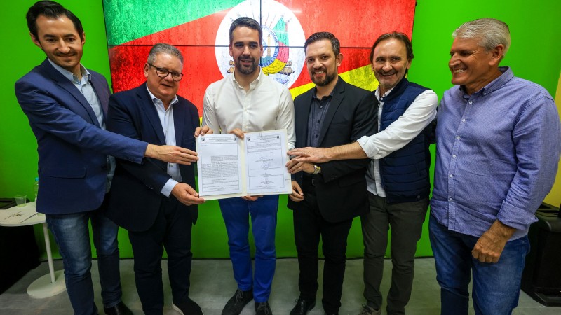 Seis homens aparecem de pé, posando de frente para a câmera. No meio, está Eduardo Leite. Eles seguram um documento assinado, que é exibido para a câmera. Todos sorriem. Ao fundo, está um painel com a bandeira do RS