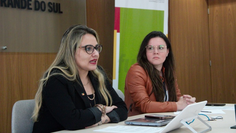 Numa sala fechada de paredes de cor marrom, as duas mulheres estão sentadas junto a uma mesa de cor branca. A foto enquadra ambas a partir de um ângulo mais à esquerda. Danielle, de casaco preto, está falando enquanto olha para a frente; Stefanie usa jaqueta marrom e está a seu lado, à direita no quadro, e olha para ela. 