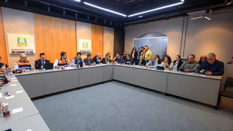 Numa sala de reuniões, os participantes estão dispostos em U, junto a uma grande mesa contínua. Leite está o meio, ao fundo do quadro, que pega a sala a partir da esquerda. Há mais de 30 pessoas presentes, e elas estão concentradas na conversa ou em seus celulares. A sala é bem iluminada, o chão e a mesa são de cor cinza e as pares são brancas. Ao fundo, atrás de Leite, há um painel que imita madeira com a logo do Estado.