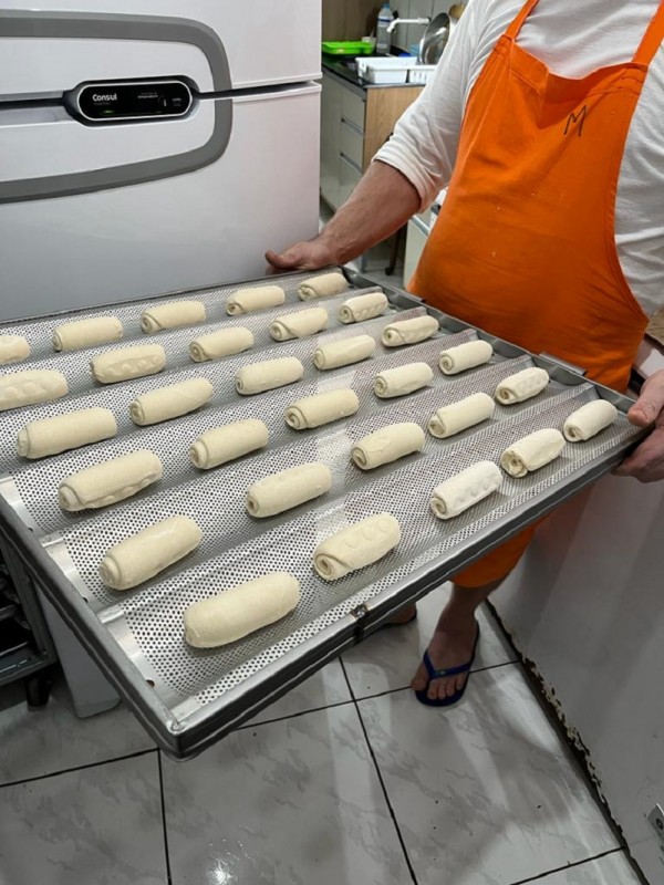 Numa cozinha, um padeiro de avental laranja exibe uma fornada de pães para assar. 