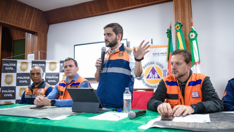 Gabriel Souza em pé atrás de uma mesa e ladeado por dois homens do lado esquerdo sentados e um do lado direito também sentado. Ele está com o microfone falando e gesticulando durante entrevista coletiva.