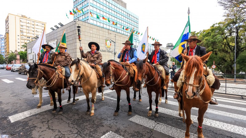 Seis homens montados a cavalo diante do Palácio Piratini. Um deles segura a tocha com a Chama Crioula e os outros bandeiras. Ao fundo, pode ser visto o prédio da Assembleia Legislativa.
