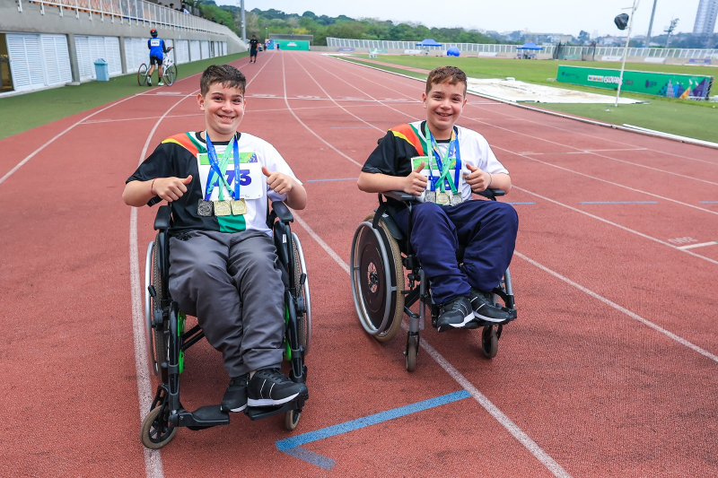 Gêmeos da delegação gaúcha das Paralimpíadas Escolares 2023 posam para a foto com suas medalhas e em cadeiras de rodas, em ambiente aberto, na pista de atletismo