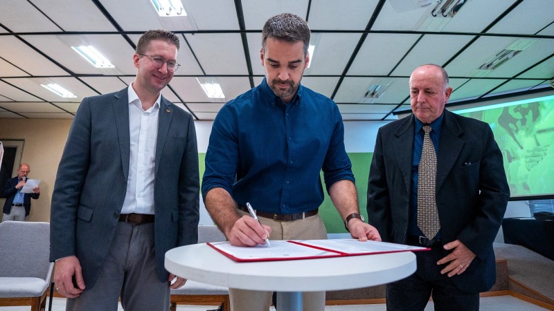 Leite ao centro em movimento de assinatura do lançamento dos editais. À sua esquerda está o secretário Beto Fantinel e há outro homem à sua direita. Os dois observam o governador assinado o documento.