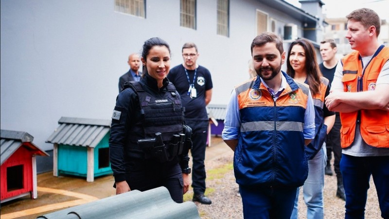 Vice-governador vendo as casinhas na penitenciária de encantado. Há uma policial do seu lado esquerdo que lhe mostra uma casinha. Há outras pessoas na imagem acompanhando Gabriel e podem ser vistas duas casinhas no canto inferior esquerdo da imagem e parte do telhado de uma delas que está diante de Gabriel.