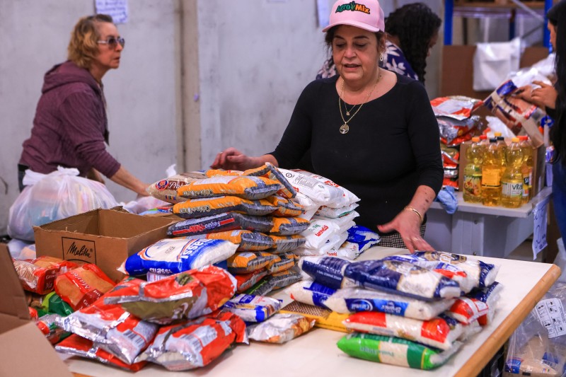 Num galpão, uma senhora de boné está junto a uma mesa com diversos pacotes de alimentos. Ao fundo, pessoas trabalham na organização dos kits.