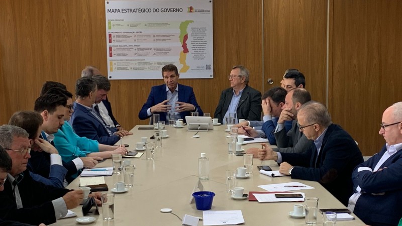 Foto em ambiente fechado, com diversas autoridades reunidas em torno de uma mesa retangular. Ao centro, está o secretário de Desenvolvimento Econômico.