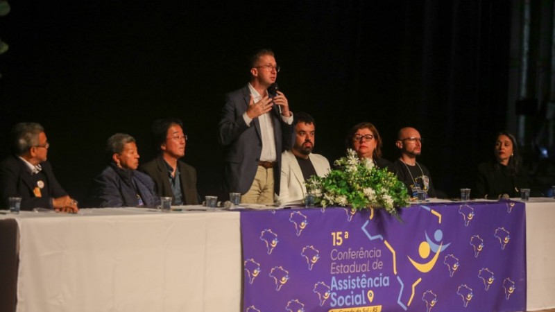 Foto com os integrantes da mesa de abertura do evento. Sete pessoas estão sentadas e, ao centro, o secretário Beto Fantinel está em pé, falando ao microfone. Em frente à mesa, há um cartaz em cor roxa identificando o evento com a inscrição "15ª Conferência Estadual de Assistência Social".