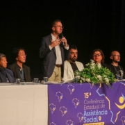 Foto com os integrantes da mesa de abertura do evento. Sete pessoas estão sentadas e, ao centro, o secretário Beto Fantinel está em pé, falando ao microfone. 