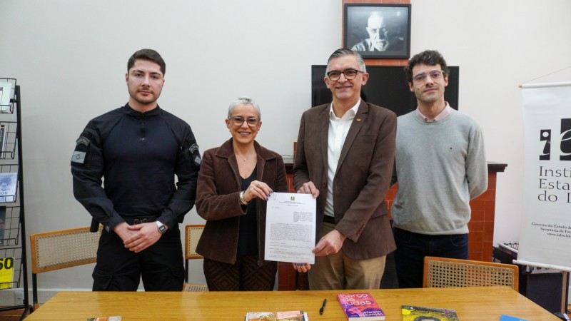 Foto posada num ambiente fechado. Na ordem da legenda, os quatro aparecem de p&eacute;, atr&aacute;s de uma mesa de madeira onde est&atilde;o depositados livros. Beatriz e Luiz seguram o documento assinado, exibindo-o para a c&acirc;mera. Ao fundo, &agrave; esquerda, h&aacute; uma prateleira com livros; &agrave; direita, aparece parte de um banner com a logo do Instituto Estadual do Livro. 