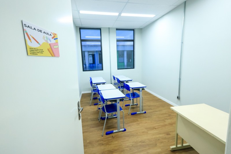 Imagem de uma das salas de aula reformadas. Sala relativamente estreita com seis mesas e seis cadeiras e uma mesa para o professor. Ao fundo, duas janelas grandes e verticais.