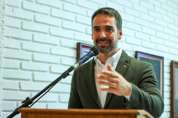 O governador Eduardo Leite, de camisa branca e paletó marrom, aparece sorrindo enquanto fala diante de microfone fixado a um pedestal. Ele gesticula com a mão direita no ar. Ao fundo, uma parede de tijolos a vista pintados de branco, na qual estão fixados alguns quadros.